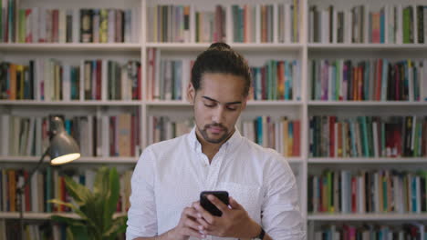 portrait-of-young-hispanic-man-student-enjoying-texting-browsing-online-using-smartphone-mobile-app-in-library-bookshelf-background