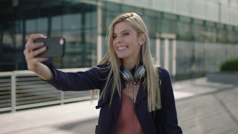 portrait-of-young-lively-blonde-business-woman-intern-smiling-posing-making-faces-taking-selfie-photo-using-smartphone-camera-in-city