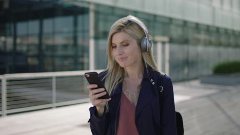 portrait-of-young-lively-blonde-business-woman-intern-happy-listening-to-music-wearing-headphones-texting-browsing-social-media-using-smartphone
