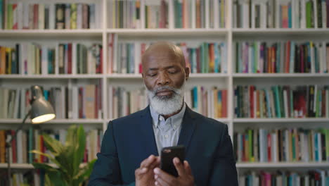 Porträt-Eines-Reifen-Afroamerikanischen-Geschäftsmann-Chefs,-Der-Ernsthaft-Aussieht-Und-SMS-Browsing-Mit-Der-Smartphone-Mobile-Networking-App-Im-Bücherregal-Hintergrund-Der-Bibliothek-Verwendet