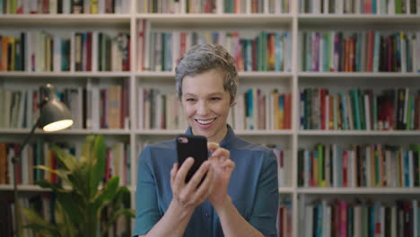 Retrato-De-Una-Mujer-Caucásica-Madura-Y-Feliz-Sonriendo-Disfrutando-De-La-Navegación-De-Mensajes-De-Texto-Usando-Tecnología-Móvil-De-Teléfono-Inteligente-En-La-Biblioteca