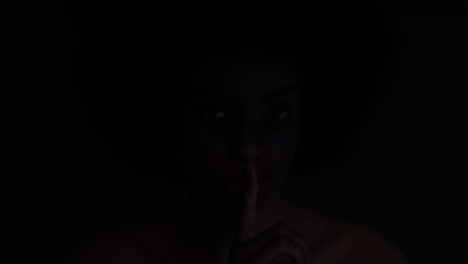 portrait-beautiful-african-american-woman-wearing-face-paint-body-art-showing-silence-gesture-mysterious-female-keeping-secret-light-flashing-in-dark-background-creative-expression-concept
