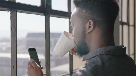 portrait-young-african-american-man-using-smartphone-drinking-coffee-at-home-browsing-messages-networking-texting-social-media-sharing-lifestyle-online-looking-out-window-in-modern-apartment-close-up