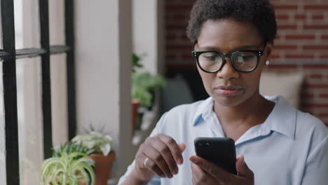 Junge-Afroamerikanische-Geschäftsfrau,-Die-Zu-Hause-Ihr-Smartphone-Nutzt-Und-Nachrichten-Durchstöbert,-Die-Online-Social-Media-Sharing-Lebensgewohnheiten-Genießt-Und-Aus-Dem-Fenster-Blickt,-SMS-Auf-Mobiltelefontechnologie-Trägt-Und-Eine-Brille-Trägt