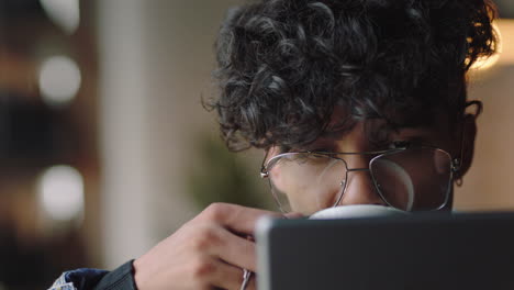 close-up-attractive-young-mixed-race-man-student-using-tablet-computer-in-cafe-browsing-online-reading-social-media-messages-enjoying-drinking-coffee-relaxing-wearing-stylish-glasses