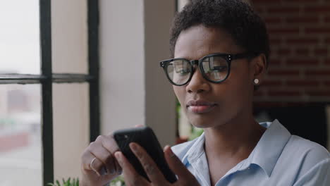 Junge-Afroamerikanische-Geschäftsfrau,-Die-Zu-Hause-Ihr-Smartphone-Nutzt-Und-Nachrichten-Durchstöbert,-Die-Online-Social-Media-Sharing-Lebensgewohnheiten-Genießt-Und-Aus-Dem-Fenster-Blickt,-SMS-Auf-Mobiltelefontechnologie-Trägt-Und-Eine-Brille-Trägt