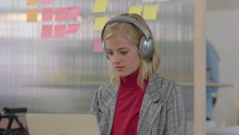 stylish-young-business-woman-using-laptop-computer-in-office-working-creative-project-typing-email-listening-to-music-wearing-headphones-looking-funny-at-camera