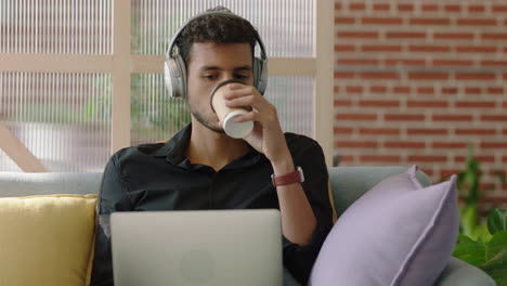 Joven-Hispano-Usando-Una-Computadora-Portátil-Navegando-Mensajes-En-Línea-Compartiendo-Comunicación-En-Red-Estudiante-Tomando-Café-Disfrutando-Escuchando-Música-En-Una-Oficina-Moderna