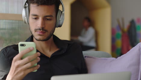 young-hispanic-man-using-laptop-computer-browsing-online-messages-sharing-network-communication-student-enjoying-listening-to-music-checking-smartphone-social-media-in-modern-office