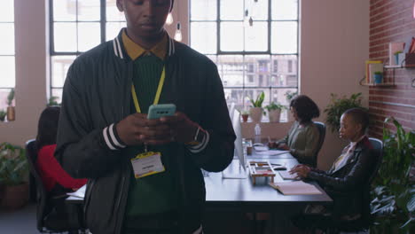 Junger-Afroamerikanischer-Student-Nutzt-Sein-Smartphone,-Um-Social-Media-Nachrichten-Zu-Durchsuchen-Und-Auf-Dem-Mobiltelefon-SMS-Zu-Schreiben,-Während-Er-An-Einem-Vielfältigen-Büroarbeitsplatz-Spaziert