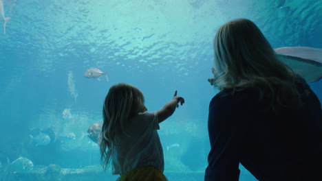 Glückliches-Mädchen-Mit-Mutter-Im-Aquarium-Und-Blick-Auf-Den-Stachelrochen,-Der-Mit-Fischen-Im-Aquarium-Schwimmt.-Kind-Beobachtet-Neugierig-Meerestiere-Und-Hat-Spaß-Daran,-Mit-Mutter-Im-Ozeanarium-Etwas-über-Das-Leben-Im-Meer-Zu-Lernen