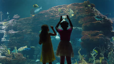 Zwei-Kleine-Mädchen-Im-Aquarium-Beobachten-Tropische-Fische-Im-Corel-Riff-Habitat,-Neugierige-Kinder,-Die-Mit-Dem-Smartphone-Im-Ozeanarium-Fotos-Von-Meerestieren-Machen-Und-Spaß-Daran-Haben,-Etwas-über-Das-Leben-Im-Meer-Zu-Lernen
