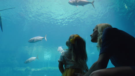 Glückliche-Mutter-Mit-Tochter-Im-Aquarium-Und-Blick-Auf-Wunderschöne-Fische,-Die-Im-Aquarium-Schwimmen.-Kleines-Mädchen-Beobachtet-Neugierig-Meerestiere-Und-Hat-Spaß-Daran,-Mit-Ihrer-Mutter-Im-Ozeanarium-Etwas-über-Das-Leben-Im-Meer-Zu-Lernen