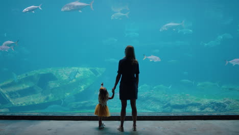 Mutter-Mit-Tochter-Im-Aquarium-Und-Blick-Auf-Wunderschöne-Fische,-Die-Im-Aquarium-Schwimmen.-Kleines-Mädchen-Beobachtet-Neugierig-Meerestiere-Und-Lernt-Mit-Ihrer-Mutter-Im-Ozeanarium-Spielerisch-Etwas-über-Das-Leben-Im-Meer