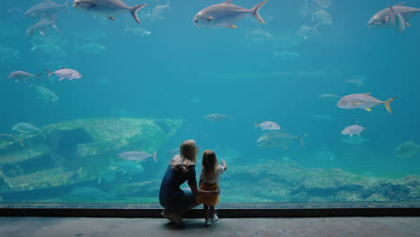 Mutter-Mit-Tochter-Im-Aquarium-Und-Blick-Auf-Wunderschöne-Fische,-Die-Im-Aquarium-Schwimmen.-Kleines-Mädchen-Beobachtet-Neugierig-Meerestiere-Und-Lernt-Mit-Ihrer-Mutter-Im-Ozeanarium-Spielerisch-Etwas-über-Das-Leben-Im-Meer