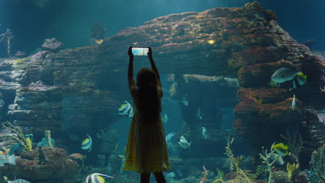 Mädchen-Benutzt-Smartphone-Im-Aquarium-Und-Fotografiert-Bunte-Fische,-Die-Mit-Meerestieren-Im-Aquarium-Schwimmen.-Neugieriges-Kind,-Das-Spaß-Daran-Hat,-Das-Meeresleben-Im-Ozeanarium-Rifflebensraum-Zu-Beobachten