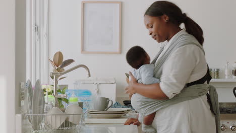 Afroamerikanische-Mutter-Hält-Ihr-Baby-Und-Arbeitet-Zu-Hause,-Wäscht-Geschirr,-Reinigt-Die-Küche-Und-Kümmert-Sich-Um-Das-Kleinkind,-Das-Hausarbeit-Erledigt-Und-Die-Mutterschaft-Genießt