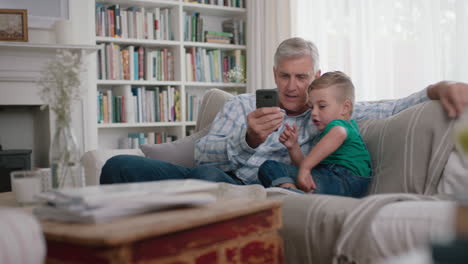 Glücklicher-Großvater,-Der-Dem-Kleinen-Jungen-Zeigt,-Wie-Man-Das-Smartphone-Benutzt,-Und-Dem-Neugierigen-Enkel-Moderne-Technologie-Beibringt,-Intelligentes-Kind,-Das-Das-Mobiltelefon-Lernt,-Während-Es-Mit-Dem-Opa-Auf-Dem-Sofa-Sitzt
