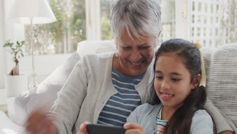 Fröhliches-Mädchen,-Das-Smartphone-Mit-Oma-Beim-Videochat-Führt,-Winkt-Der-Familie-Zu-Und-Teilt-Ihr-Urlaubswochenende-Mit-Großmutter,-Die-Auf-Dem-Handy-Chattet-Und-Sich-Zu-Hause-Mit-Enkelin-In-4K-Entspannt