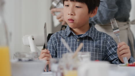 Feliz-Niño-Asiático-Comiendo-Gofres-Frescos-Para-El-Desayuno-Disfrutando-De-Una-Deliciosa-Comida-Casera-Con-La-Familia-En-La-Cocina-De-Casa-4k