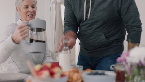 Glückliches-älteres-Paar,-Das-Gemeinsam-Frühstückt,-Ehemann-Auf-Der-Suche-Nach-Milch-Im-Kühlschrank,-Sitzend-Mit-Frau-Am-Tisch-Und-Gemeinsames-Morgenessen-In-Der-Küche,-Tag-Im-Leben,-4K-Videos