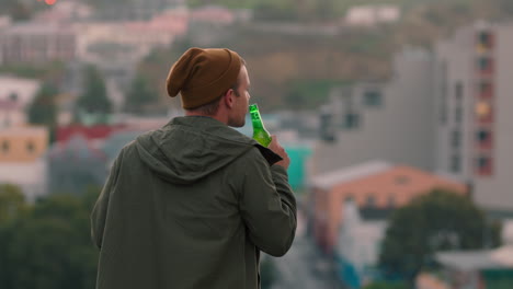 young-caucasian-man-drinking-alcohol-on-rooftop-enjoying-cold-refreshing-beer-looking-at-city-skyline