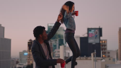 happy-multiracial-couple-enjoying-rooftop-date-young-woman-walking-on-edge-drinking-alcohol-enjoying-carefree-weekend-lifestyle-in-city-at-sunset