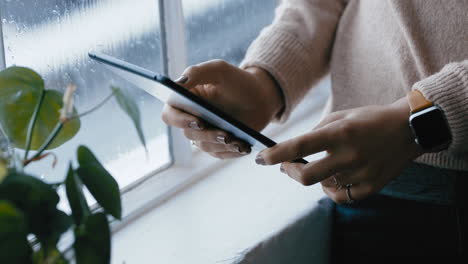 Nahaufnahme-Von-Frauenhänden,-Die-Einen-Digitalen-Tablet-Computer-Nutzen-Und-Online-Nachrichten-Durchsuchen,-Soziale-Medien-Lesen-Und-Ein-Mobiles-Touchscreen-Gerät-Genießen,-Das-Am-Fenster-Steht-Und-Sich-An-Einem-Kalten,-Regnerischen-Tag-Zu-Hause-Entspannt