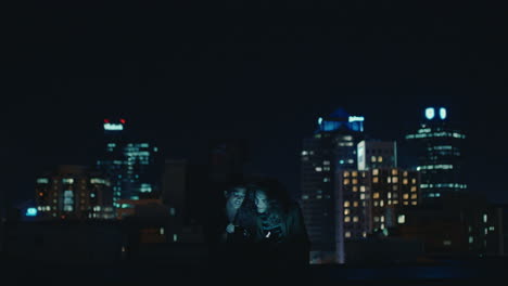 friends-using-smartphone-mobile-technology-hanging-out-on-rooftop-at-night-enjoying-weekend-chatting-sharing-social-media-messages-in-urban-city-skyline-background