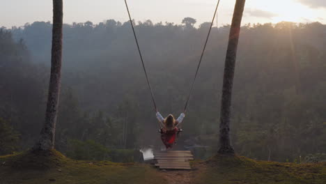 aerial-view-woman-swinging-over-tropical-rainforest-at-sunrise-sitting-on-swing-with-view-of-river-enjoying-having-fun-on-holiday-travel-freedom