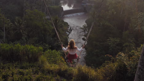 top-view-woman-swinging-over-tropical-rainforest-at-sunrise-sitting-on-swing-with-view-of-river-enjoying-having-fun-on-holiday-travel-freedom