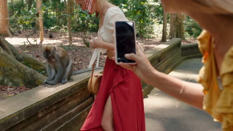 Mujer-Turista-Usando-Un-Teléfono-Inteligente-Tomando-Una-Foto-De-Un-Amigo-Posando-Con-Un-Mono-En-Un-Zoológico-De-Vida-Silvestre-Divirtiéndose-Compartiendo-Aventuras-De-Viaje