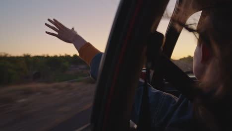 woman-in-car-holding-hand-out-window-feeling-wind-blowing-through-fingers-driving-in-countryside-on-road-trip-travelling-for-summer-vacation-enjoying-freedom-on-the-road-at-sunrise