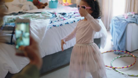 happy-little-girl-playing-dress-up-dancing-in-bedroom-with-mother-taking-photo-using-smartphone-sharing-on-social-media