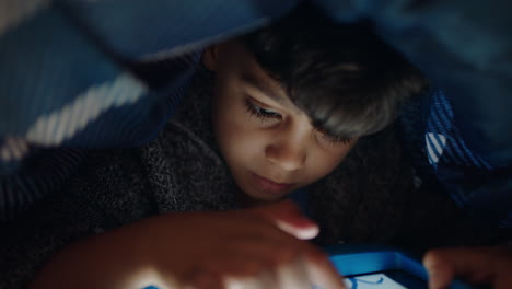 Niedlicher-Kleiner-Junge,-Der-Unter-Einer-Decke-Einen-Digitalen-Tablet-Computer-Benutzt-Und-Es-Genießt,-Auf-Der-Touchscreen-Technologie-Zu-Zeichnen,-Spiele-Zu-Spielen-Und-Vor-Dem-Schlafengehen-Spaß-Zu-Haben