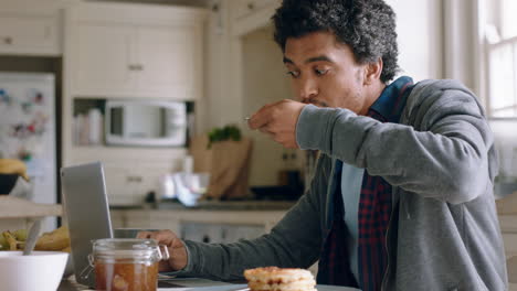 happy-mixed-race-man-using-laptop-computer-working-in-kitchen-eating-waffles-for-breakfast-browsing-online-enjoying-relaxing-morning-at-home