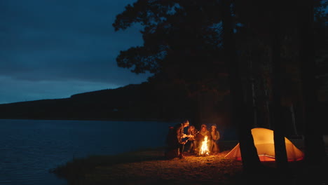 Eine-Gruppe-Von-Freunden-Sitzt-Am-Lagerfeuer-Und-Röstet-Marshmallows,-Zeltet-Im-Wald-Am-See-Und-Unterhält-Sich-Nachts,-Teilt-Wärme-Und-Genießt-Outdoor-Abenteuer-In-4k