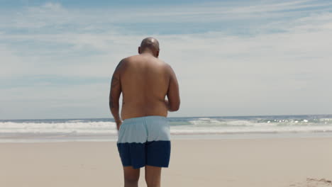 Junger-Afroamerikanischer-Mann,-Der-Am-Strand-Spaziert-Und-Den-Warmen-Sommertag-Genießt-Und-Sich-Zum-Schwimmen-Vorbereitet,-Rückansicht-4k
