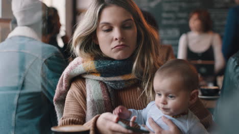 Madre-Joven-Con-Bebé-En-La-Cafetería-Usando-Un-Teléfono-Inteligente-Tomando-Café-Relajándose-En-Un-Restaurante-Concurrido-Disfrutando-De-La-Maternidad