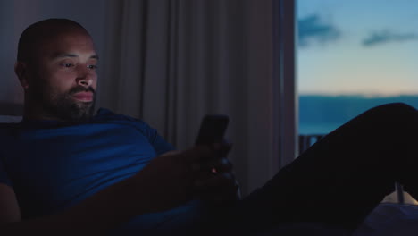 young-man-using-smartphone-lying-in-bed-texting-browsing-social-media-messages-enjoying-online-entertainment-relaxing-in-apartment-at-sunset