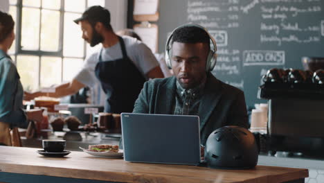 Junger-Afroamerikanischer-Mann-Benutzt-Laptop-Im-Café,-Surft-Online-Und-Hört-Musik,-Trägt-Kopfhörer-Und-Genießt-Mobile-Computertechnologie