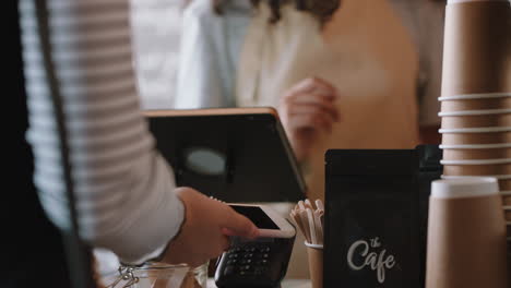 Glückliche-Barista-Frau,-Die-Den-Kunden-Bedient-Und-Mit-Dem-Smartphone-Kontaktlos-Bezahlt-Und-Geld-Ausgibt,-Um-Kaffee-Im-Café-Zu-Kaufen