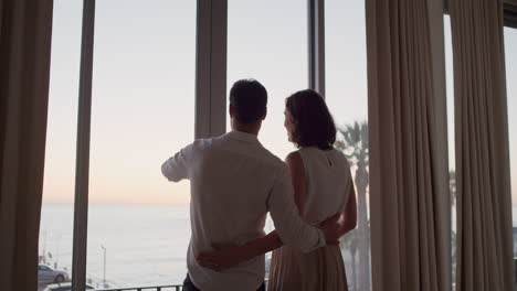 happy-travel-couple-arriving-in-hotel-room-on-vacation-looking-out-window-young-man-photographing-view-enjoying-romantic-holiday-with-wife