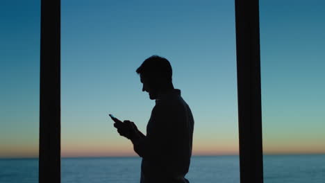 Junger-Mann-Benutzt-Smartphone-Im-Hotelzimmer-Und-Schreibt-SMS,-Teilt-Seinen-Urlaubslebensstil-In-Den-Sozialen-Medien-Und-Genießt-Den-Blick-Auf-Das-Meer-Bei-Sonnenuntergang