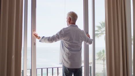 Glücklicher-Alter-Mann-Tanzt-Und-Genießt-Die-Schöne-Aussicht-Auf-Das-Meer-Im-Hotelzimmer,-Um-Einen-Erfolgreichen-Ruhestandsurlaub-Zu-Feiern