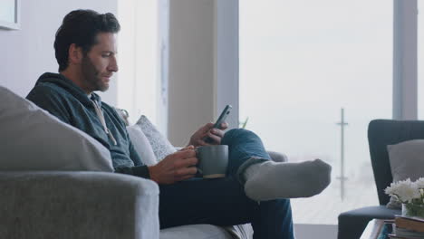 young-man-using-smartphone-browsing-online-reading-social-media-messages-enjoying-comfortable-lifestyle-drinking-coffee-relaxing-on-couch-at-home