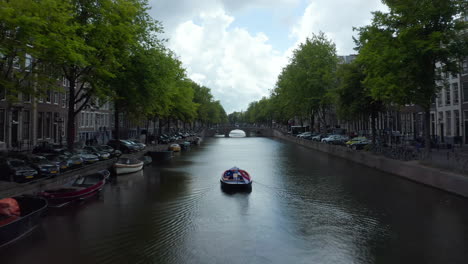 Canal-De-Amsterdam-Con-Barco-Y-Bandera-Holandesa-Ondeando-En-El-Viento,-Antena-Delantera