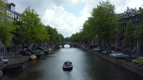 Establecedor-Sobre-El-Canal-De-Amsterdam-Con-Barco-Y-Bandera-Holandesa-Ondeando-En-El-Viento,-Antena-Hacia-Atrás