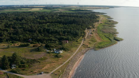 Aerial-shot-of-sea-coast.-Cottages-at-forest-and-walkway-along-shore.-Wind-park-providing-green-sustainable-energy-in-background.-Denmark