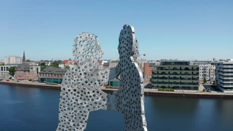 Orbit-shot-around-top-part-of-Molecule-Man-aluminium-work-of-art.-Buildings-on-Spree-river-waterfront-in-background.-Berlin,-Germany